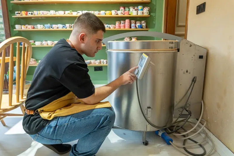 Man in pottery studio using digital control panel on a kiln, surrounded by colorful ceramics