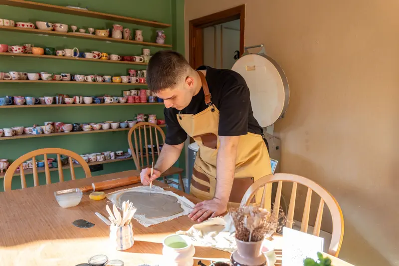 A man wearing an apron creates pottery in a sunlight-filled art studio with shelves of clay pots