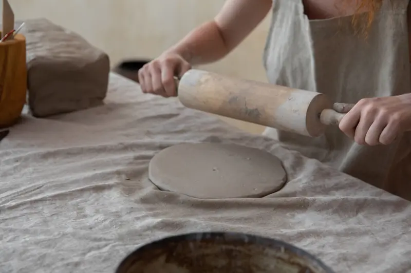 Crop anonymous female artisan in apron rolling out clay slab on table while creating in light pottery