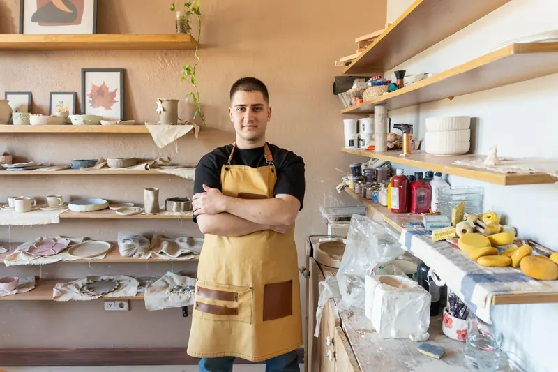 Young potter in apron poses confidently in a well-organized ceramics studio filled with pottery and tools